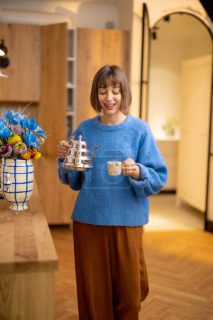 Téléchargez les photos : La jeune femme prépare une boisson au café, debout avec une bouilloire et une tasse à l'intérieur de la cuisine moderne. Concept de bien-être et mode de vie domestique - en image libre de droit