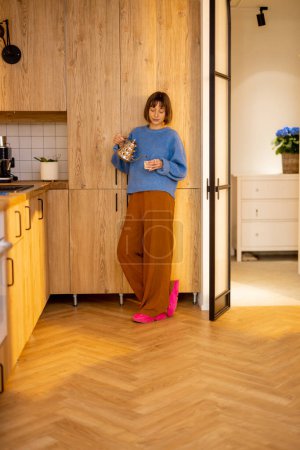 Foto de Mujer joven pasa un tiempo libre de pie con un hervidor de agua y una taza en la cocina de su apartamento estudio moderno. Concepto de estilo de vida y bienestar doméstico - Imagen libre de derechos