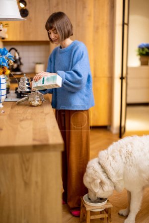 Téléchargez les photos : Jeune femme fait un petit déjeuner avec du yaourt et du granola tout en se tenant avec son chien mignon sur la cuisine à la maison. Style de vie domestique et amitié avec les animaux concept - en image libre de droit