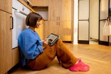 Photo for Woman designing kitchen interior on a digital tablet while sitting on a kitchen floor. Concept of designing interiors using digital devices and work from home - Royalty Free Image