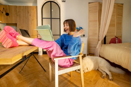 Photo for Young woman sits relaxed with legs on table while having online call on a digital tablet at modern studio apartment with her dog resting behind - Royalty Free Image