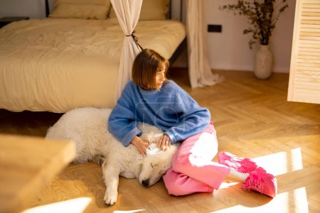 Foto de Mujer joven cuida a su lindo perro blanco mientras están acostados juntos en el suelo en casa. Tiempo libre y amistad con el concepto de perro - Imagen libre de derechos