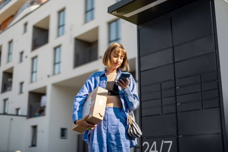 Photo for Young woman using smart phone while standing with a parcel delivered with post office machine with automatic lockers. New technologies in delivery service, self picking - Royalty Free Image