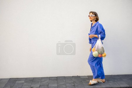 Téléchargez les photos : Femme en pyjama bleu marche avec sac en filet plein de fruits frais sur fond de mur blanc à l'extérieur. Concept de durabilité et mode de vie moderne - en image libre de droit