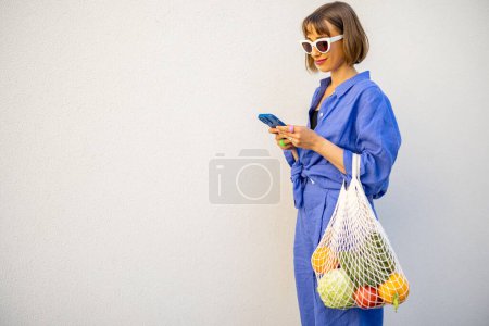 Téléchargez les photos : Femme en pyjama bleu marche avec sac en filet plein de fruits frais sur fond de mur blanc à l'extérieur. Concept de durabilité et mode de vie moderne - en image libre de droit