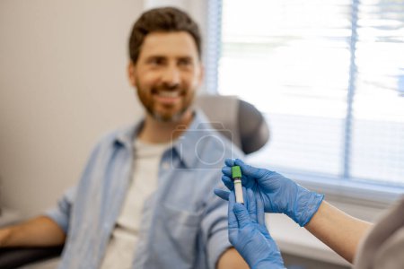 Foto de Asistente de laboratorio sostiene tubo con sangre para el análisis, paciente varón feliz en el fondo, se centran en un tubo - Imagen libre de derechos