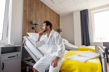 Téléchargez les photos : Homme mûr réfléchi assis sur le lit dans une salle d'un hôpital moderne, se reposant pendant la réadaptation après un traitement médical - en image libre de droit