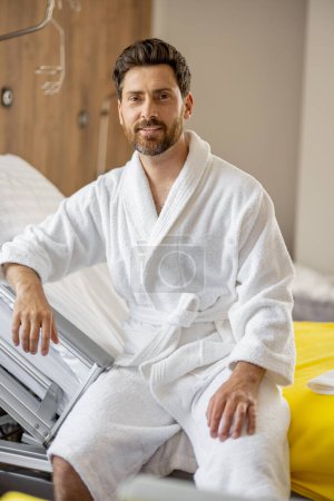 Téléchargez les photos : Portrait d'un homme adulte gai assis sur le lit dans une salle d'un hôpital moderne, se reposant pendant la réadaptation après un traitement médical - en image libre de droit