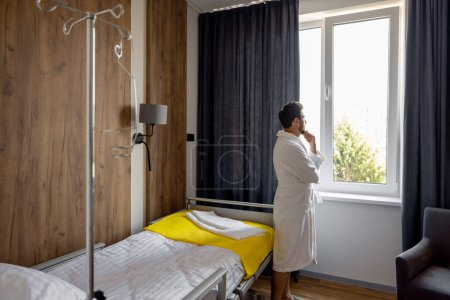 Photo for Man looks out the window, standing in medical ward alone. Medical treatment, rehabilitation after surgery, comfortable suite concept - Royalty Free Image
