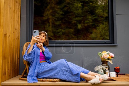 Foto de Mujer joven descansando en la terraza de madera de una casa en la naturaleza, hablando por teléfono y disfrutando de las vacaciones. Escapar a la naturaleza, la calma y el concepto de relajación - Imagen libre de derechos
