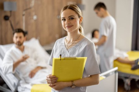 Photo for Portrait of a young female nurse standing in medical ward with patients resting on background. Nursing and medical support concept - Royalty Free Image