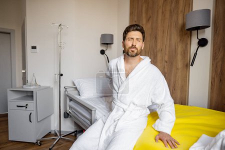Foto de Hombre sintiendo dolor de espalda mientras está sentado en la cama en la sala médica. Paciente adulto medio con alguna enfermedad en tratamiento médico en la clínica moderna - Imagen libre de derechos