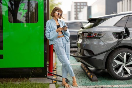 Téléchargez les photos : Une jeune femme se tient debout avec un téléphone intelligent, attendant que sa voiture électrique se recharge sur une borne de recharge publique à l'extérieur. Concept de mode de vie moderne et d'énergie verte pour les transports - en image libre de droit