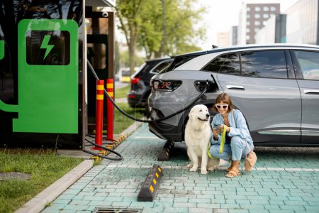 Foto de Mujer joven con un enorme perro blanco esperando que el coche eléctrico se cargue en una estación pública al aire libre. Concepto de coches EV y amistad con mascotas - Imagen libre de derechos
