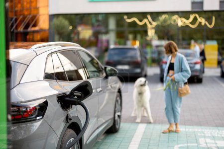 Young woman with a huge white dog waiting for electric car to be charged on a public station outdoors. Concept of EV cars and friendship with pets