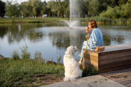 Téléchargez les photos : Jeune femme mange de la crème glacée tout en étant assis avec son chien blanc mignon près du lac dans le parc. Amitié avec les animaux domestiques et passer du temps libre ensemble - en image libre de droit