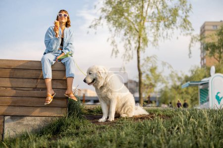 Téléchargez les photos : Jeune femme mangeant de la crème glacée tout en étant assis sur le banc avec son chien blanc mignon dans le parc. Passer du temps libre ensemble - en image libre de droit