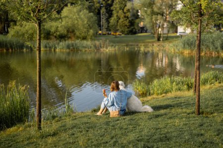 Téléchargez les photos : Jeune femme mange de la crème glacée tout en étant assis avec son chien blanc mignon près du lac dans le parc. Vue large. Amitié avec les animaux domestiques et passer du temps libre ensemble - en image libre de droit