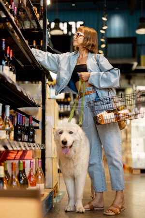 Photo for Young woman choosing wine to buy, visiting wine shop with a huge white dog. Concept of alcohol buying and pet-friendly shops - Royalty Free Image