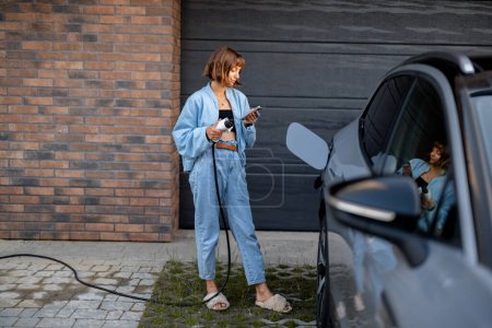 Foto de Mujer joven enchufes pistola de carga en su coche, mientras que de pie con un teléfono inteligente cerca de las puertas de garaje de su casa. Concepto de energía verde y barata de la carga de vehículos domésticos - Imagen libre de derechos