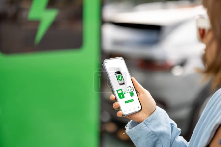 Téléchargez les photos : Femme tient un smartphone avec une application en cours d'exécution pour la charge du véhicule, charge de voiture électrique en arrière-plan, gros plan - en image libre de droit