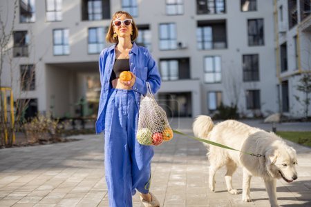 Photo for Young woman in blue pajamas walks with her dog and carry mesh bag full of fresh fruits and vegetables at inner yard of apartment building. Sustainable and modern lifestyle concept - Royalty Free Image