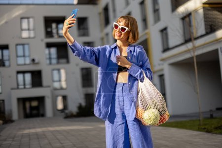 Téléchargez les photos : Happy woman parle au téléphone en ligne tout en marchant à la maison avec un sac en filet plein de fruits et légumes frais à la cour intérieure de l'immeuble - en image libre de droit