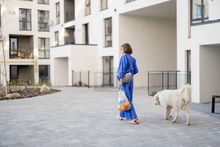 Foto de Mujer joven en pijama azul pasea con su perro y lleva una bolsa de malla llena de frutas y verduras frescas en el patio interior del edificio de apartamentos. Concepto de estilo de vida sostenible y moderno - Imagen libre de derechos