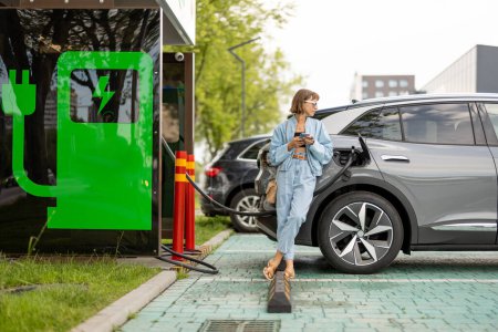 Foto de Mujer joven se para con un teléfono inteligente, esperando a que su coche eléctrico para cargar en una estación de carga pública al aire libre. Concepto de estilo de vida moderno y energía verde para el transporte - Imagen libre de derechos