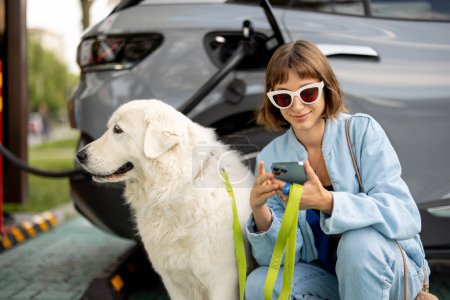 Foto de Mujer joven con un enorme perro blanco esperando que el coche eléctrico se cargue en una estación pública al aire libre. Concepto de coches EV y amistad con mascotas - Imagen libre de derechos