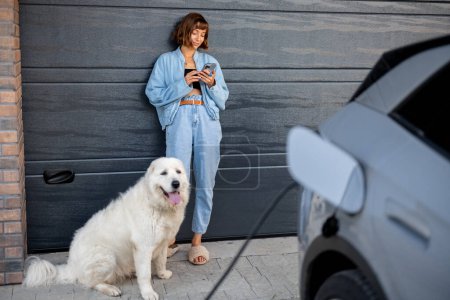 Téléchargez les photos : Femme utilise son téléphone appuyé contre la porte de garage de sa maison, un chien blanc à ses côtés. Une voiture électrique se charge au premier plan - en image libre de droit