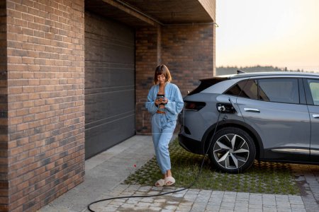 Téléchargez les photos : Jeune femme utilisant un téléphone intelligent tout en rechargeant sa voiture électrique près du garage de sa maison. Concept de mode de vie moderne et de durabilité - en image libre de droit