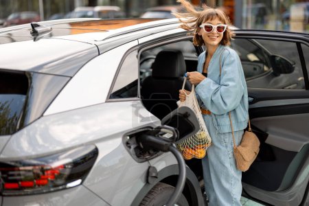 Photo for Woman puts purchased products on the back seat of her electric car, coming from the supermarket to the charging station - Royalty Free Image