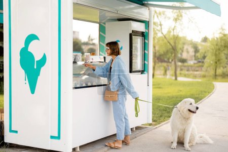 Foto de Mujer joven eligiendo helado para comprar en una hermosa tienda mientras pasea con su perro en un parque. Pasar tiempo de verano junto con mascotas al aire libre - Imagen libre de derechos
