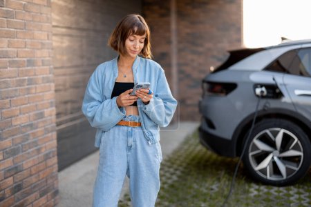 Photo for Young woman using smart phone while charging her electric car near garage of her house. Concept of modern lifestyle and sustainability - Royalty Free Image