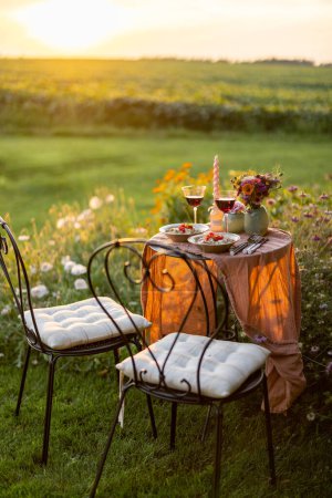 Photo for Beautiful green lawn with flowers and dining table for two during the sunset. Romantic picnic on nature - Royalty Free Image