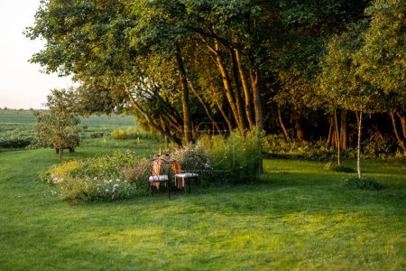 Foto de Beautiful green lawn with flowers and dining table for two near the forest during the sunset. Romantic picnic on nature and beauty of nature concept - Imagen libre de derechos