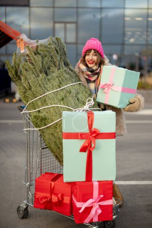 Foto de Retrato de una joven mujer alegre con carrito de compras lleno de regalo y árbol de Navidad cerca del centro comercial al aire libre. Vacaciones de invierno concepto de compras - Imagen libre de derechos