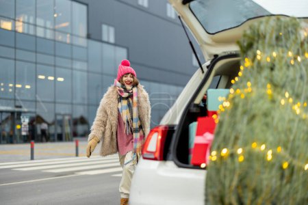 Foto de Joven mujer alegre en ropa de invierno corre cerca del centro comercial con un coche lleno de regalos y árbol de Navidad en un primer plano - Imagen libre de derechos