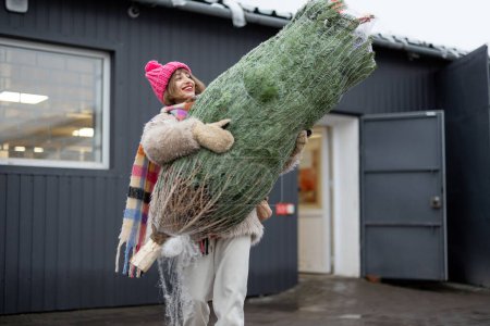 Foto de Mujer joven lleva el árbol de Navidad envuelto mientras sale de la oficina de correos entregado desde la tienda en línea. Concepto de entrega y compras en línea en vacaciones de invierno - Imagen libre de derechos