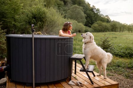 Foto de Mujer bañándose en bañera de hidromasaje al aire libre mientras descansa con su lindo perro en casa en las montañas. Concepto de recreación y pasar tiempo libre con mascotas en la naturaleza - Imagen libre de derechos
