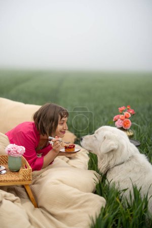 Foto de Mujer joven disfruta de la mañana, acostada en la cama con su lindo perro y comiendo desayuno dulce en el campo verde al aire libre - Imagen libre de derechos