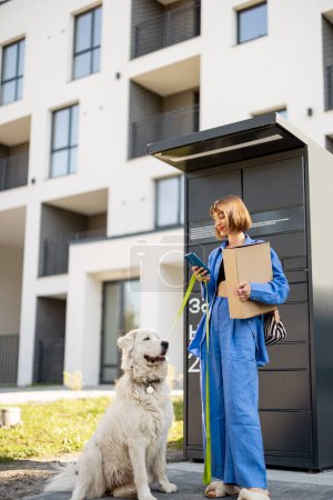 Photo for Young woman picks up parcels from automatic post office machine, standing with her dog near apartment building. Concept of fast delivery and urban lifestyle - Royalty Free Image