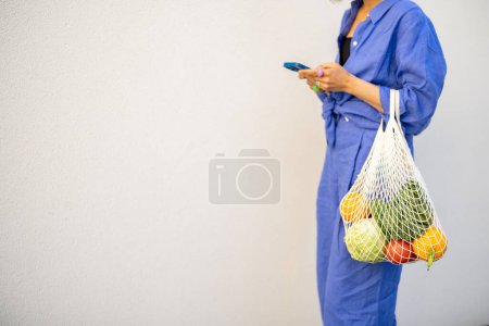 Photo for Woman in blue stands with mesh bag full of fresh fruits on white wall background outdoors. Close-up on bag - Royalty Free Image