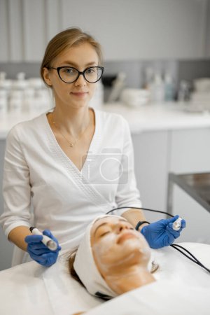 Photo for Young beautician doing microcurrent therapy on a clients face in beauty salon. Concept of cosmetology procedures for facial care. Portrait of a medical cosmetologist at work - Royalty Free Image
