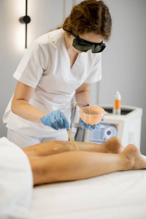 Foto de Trabajador joven de un salón de belleza durante el procedimiento de depilación en las piernas de una mujer. Concepto de depilación y procedimientos de belleza - Imagen libre de derechos