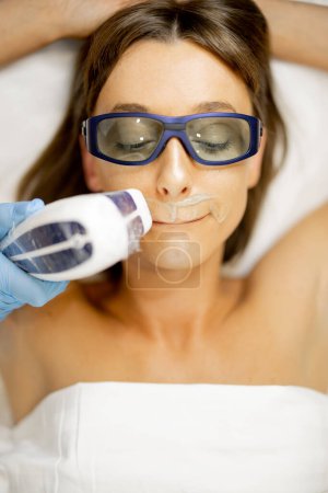 Téléchargez les photos : Jeune femme en glasse lors de la procédure d'épilation sur la lèvre supérieure au salon de beauté, vue rapprochée d'en haut. Concept d'épilation laser et de soins de la peau - en image libre de droit