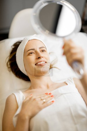 Téléchargez les photos : Femme heureuse regarde dans le miroir sur son visage après les procédures de beauté tout en étant allongé au salon de beauté. Concept de beauté et de soin du visage - en image libre de droit