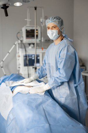 Foto de Retrato de un cirujano en uniforme estéril listo para una operación de pie cerca de un paciente acostado en quirófano. Concepto de esterilidad durante el tratamiento quirúrgico y el procedimiento invasivo - Imagen libre de derechos