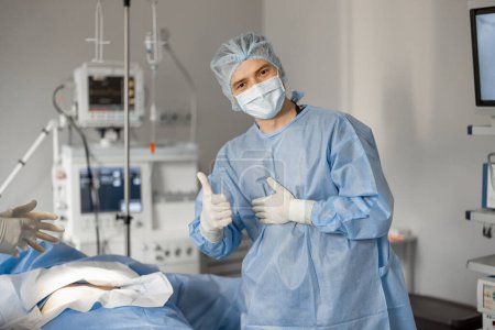 Foto de Retrato de un cirujano feliz en uniforme estéril listo para una operación de pie cerca de un paciente acostado en quirófano. Concepto de esterilidad durante el tratamiento quirúrgico y el procedimiento invasivo - Imagen libre de derechos